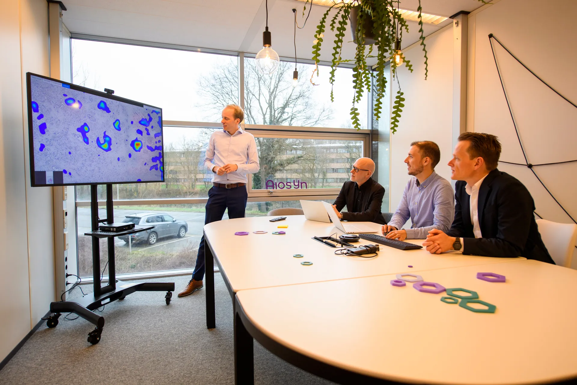 Four people around a table at Aiosyn, looking at a large screen showing a tissue section with an AI heatmap overlay. Purple and green hexagon models sit on the table.