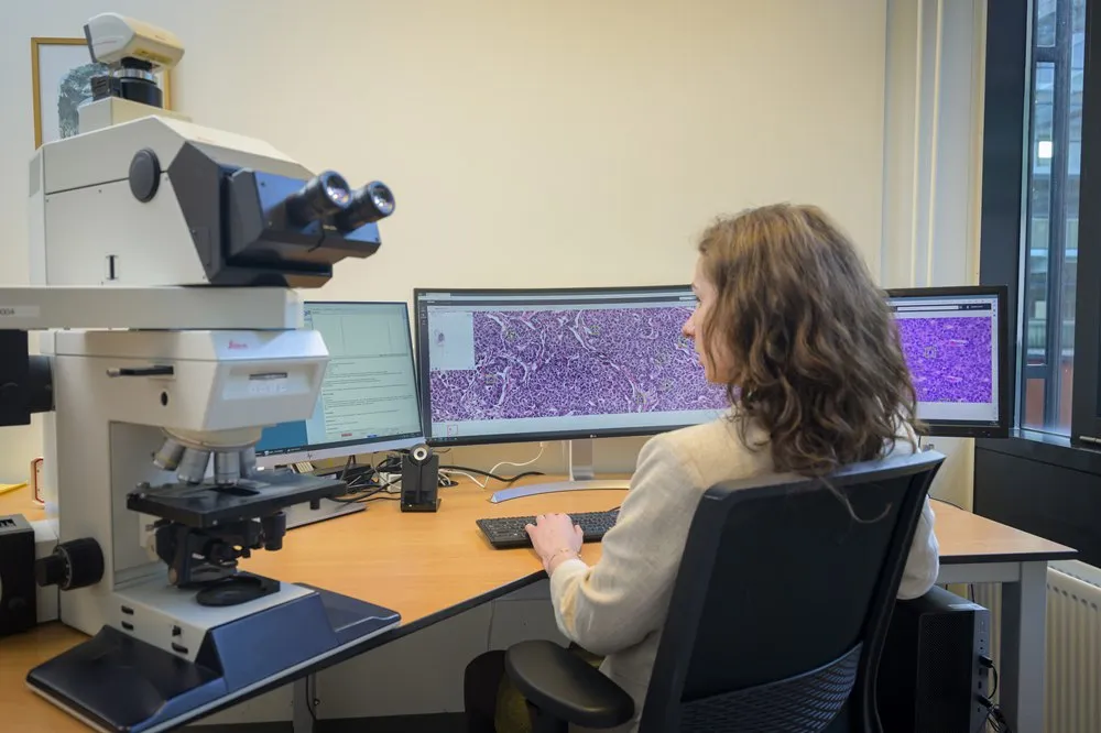 A pathologist at her workstation, reading a whole-slide image with Aiosyn's mitosis detections marked on it, the microscope still on the bench beside her.